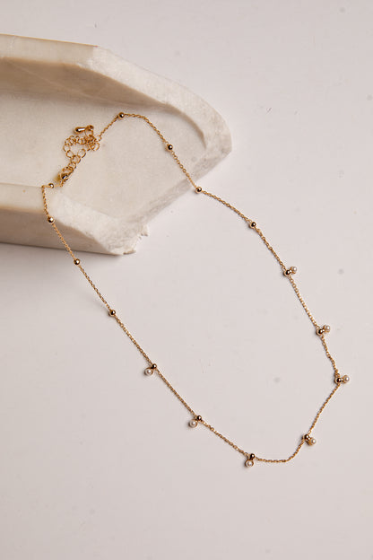 A delicate gold chain necklace with small bead accents and tiny clear stones, displayed on a light beige surface next to a textured cream stone prop.