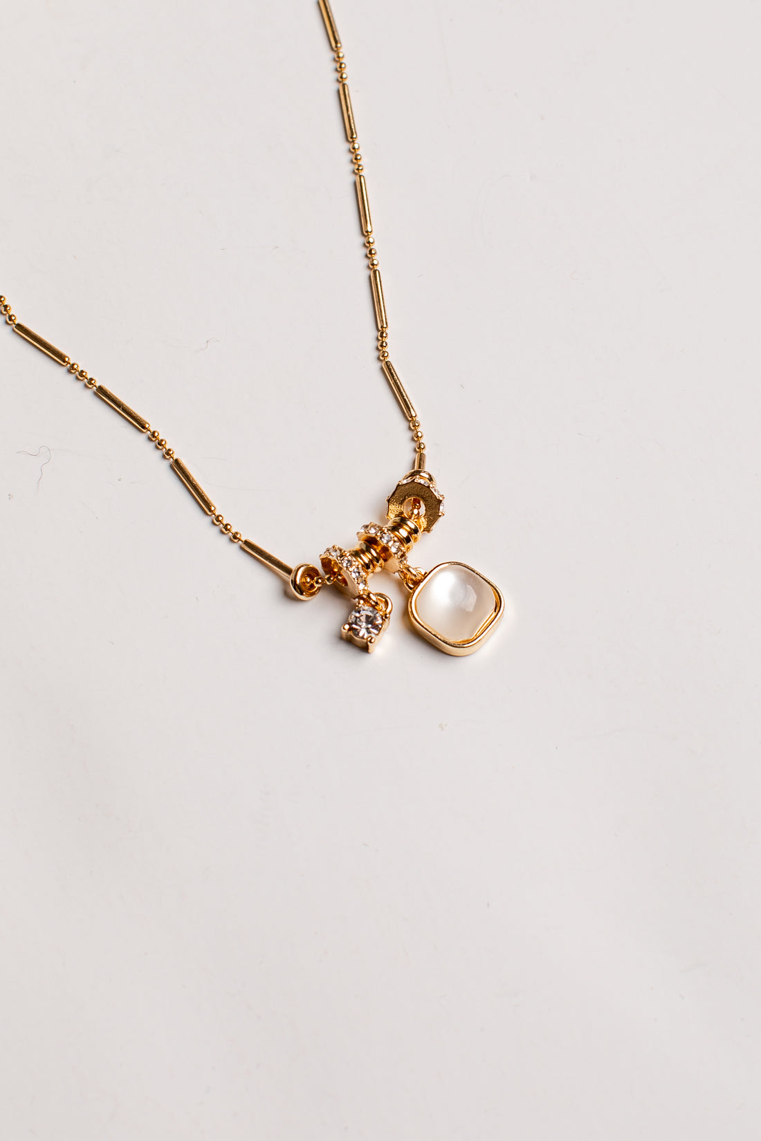 A gold chain necklace featuring a multi-charm pendant with a large square cream-colored stone, a small faceted gold bead, and a tiny clear crystal charm, displayed on a clean off-white background.