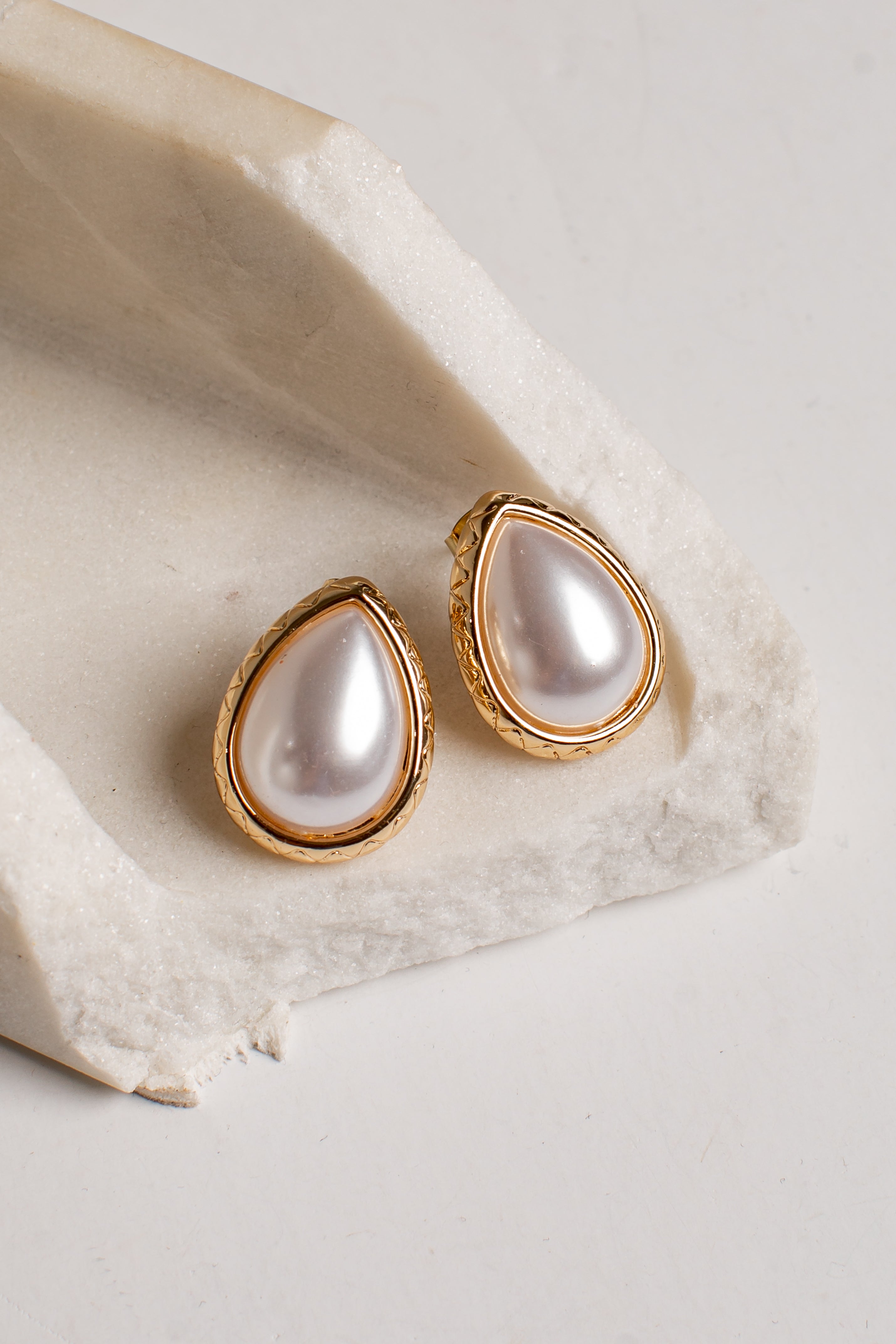 A pair of gold teardrop stud earrings with lustrous white pearl centers, displayed on a textured white stone slab against a light neutral background.
