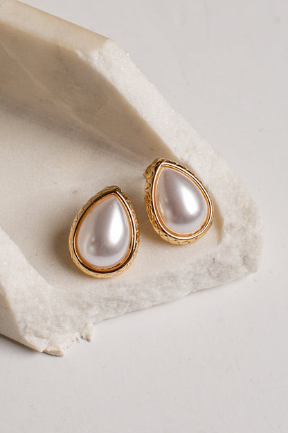 A pair of gold teardrop stud earrings with lustrous white pearl centers, displayed on a textured white stone slab against a light neutral background.