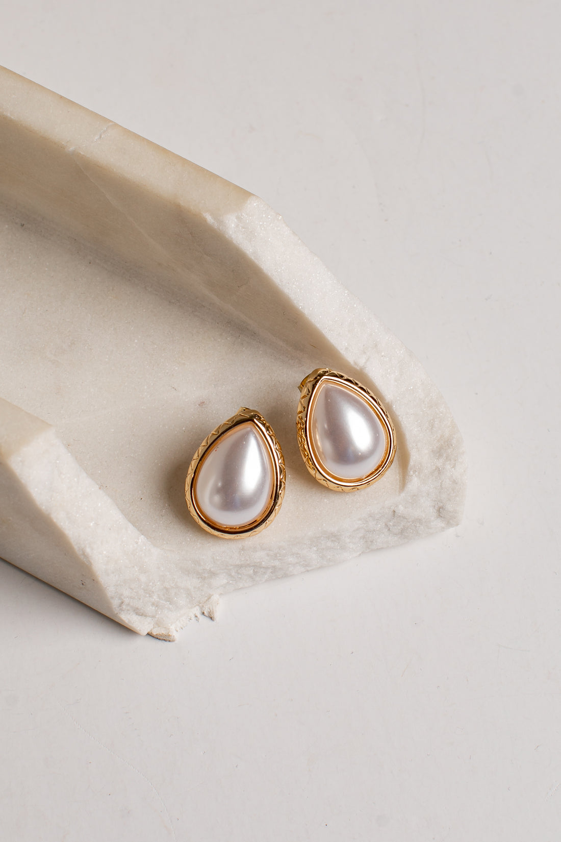 Teardrop-shaped gold stud earrings with white faux pearls, displayed on a textured beige stone riser against a light gray background.
