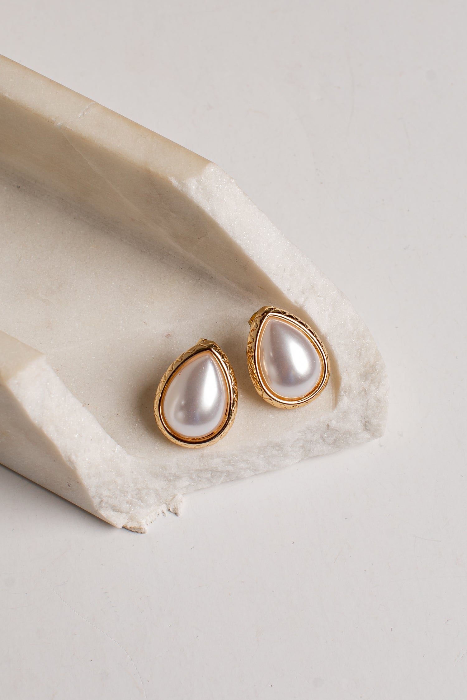 Teardrop-shaped gold stud earrings with white faux pearls, displayed on a textured beige stone riser against a light gray background.