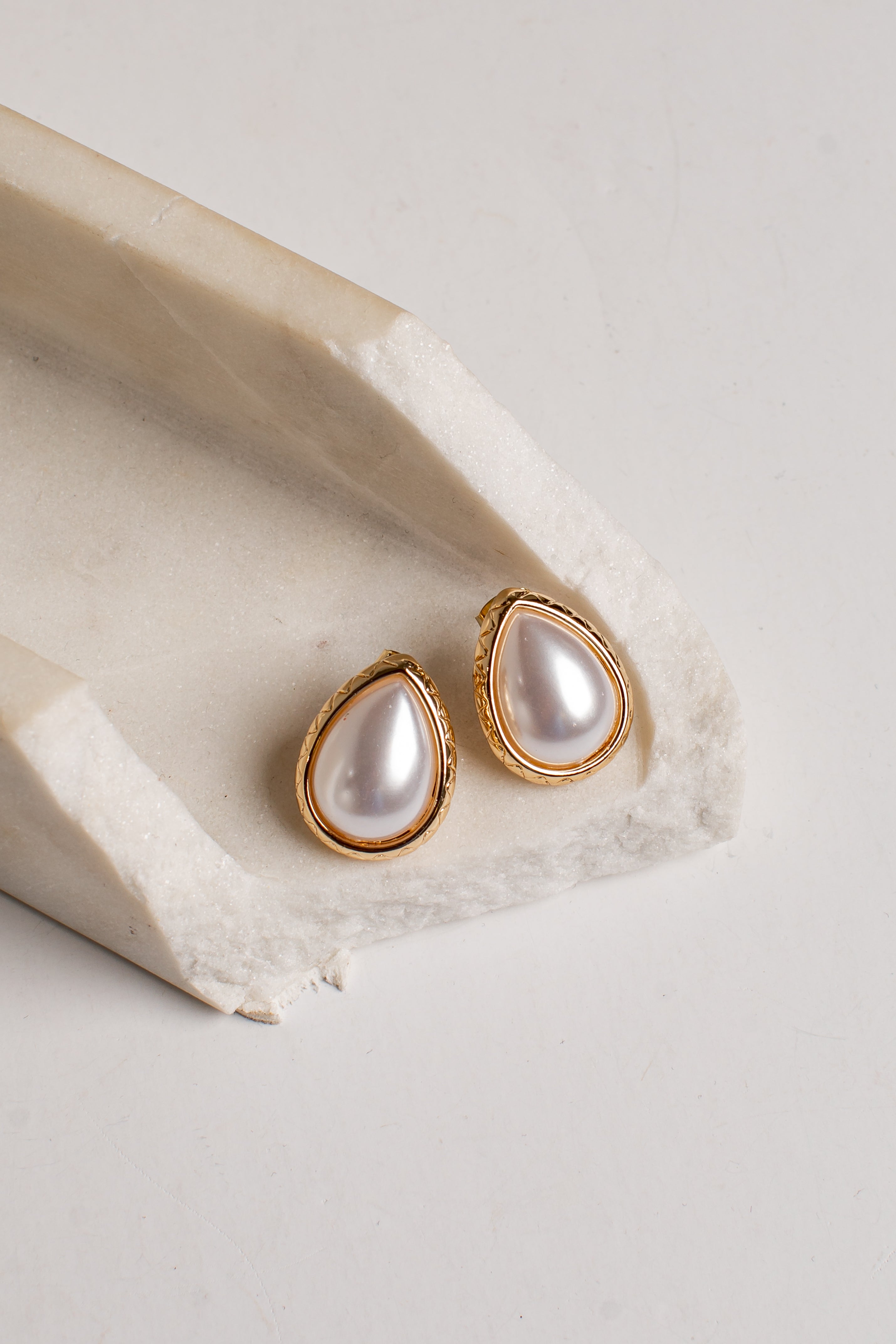 Teardrop-shaped gold stud earrings with white faux pearls, displayed on a textured beige stone riser against a light gray background.