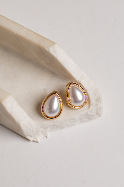 Teardrop-shaped gold stud earrings with white faux pearls, displayed on a textured beige stone riser against a light gray background.