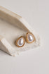 Teardrop-shaped gold stud earrings with white faux pearls, displayed on a textured beige stone riser against a light gray background.