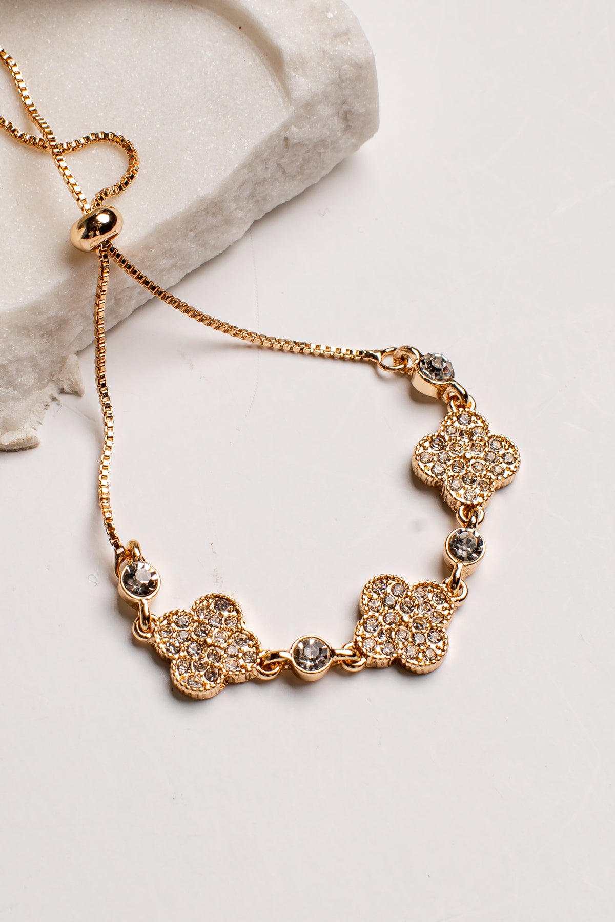 A gold-plated chain necklace with four pavé-set floral clover motifs and small clear rhinestone accents, displayed on a light surface next to a white textured stone prop.