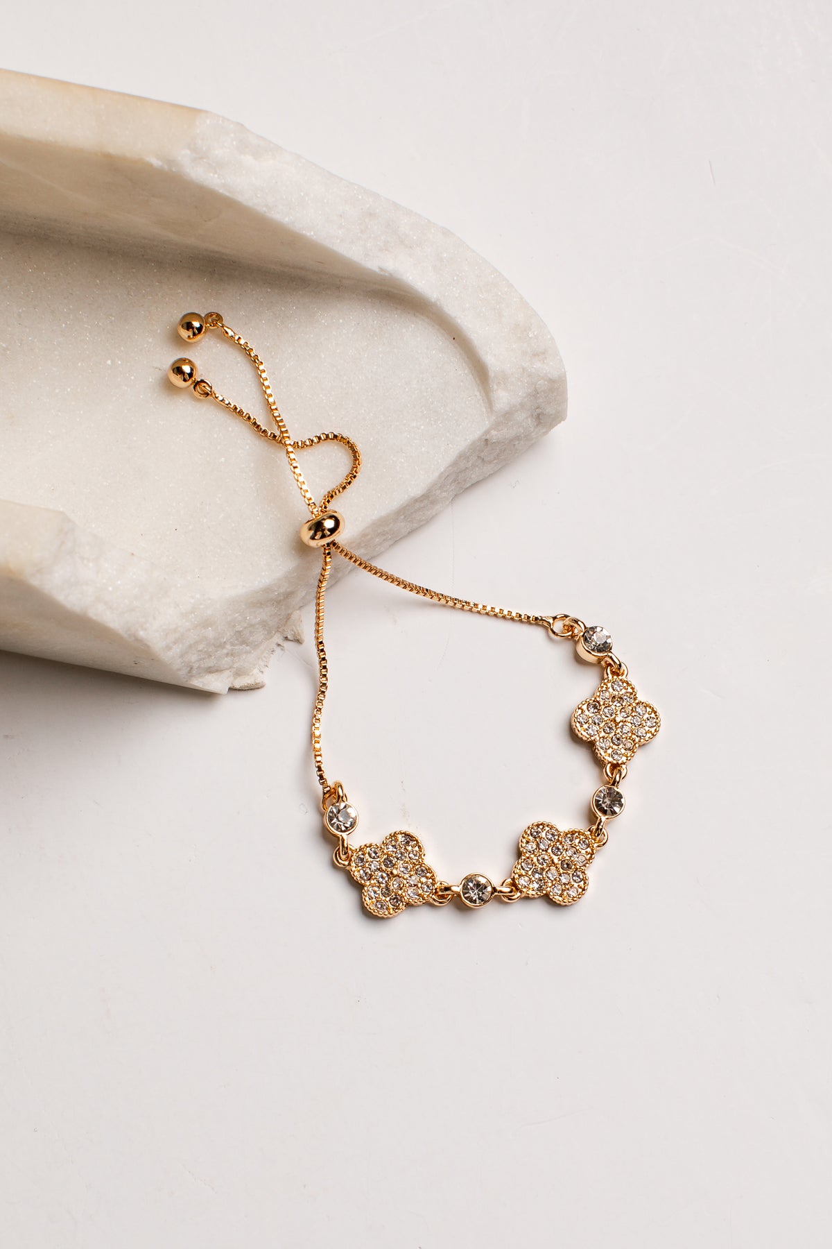A gold-toned adjustable chain bracelet with four flower-shaped charms set with clear stones, displayed on a light beige stone slab against a white background.