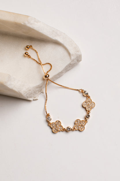 A gold-toned adjustable chain bracelet with four flower-shaped charms set with clear stones, displayed on a light beige stone slab against a white background.