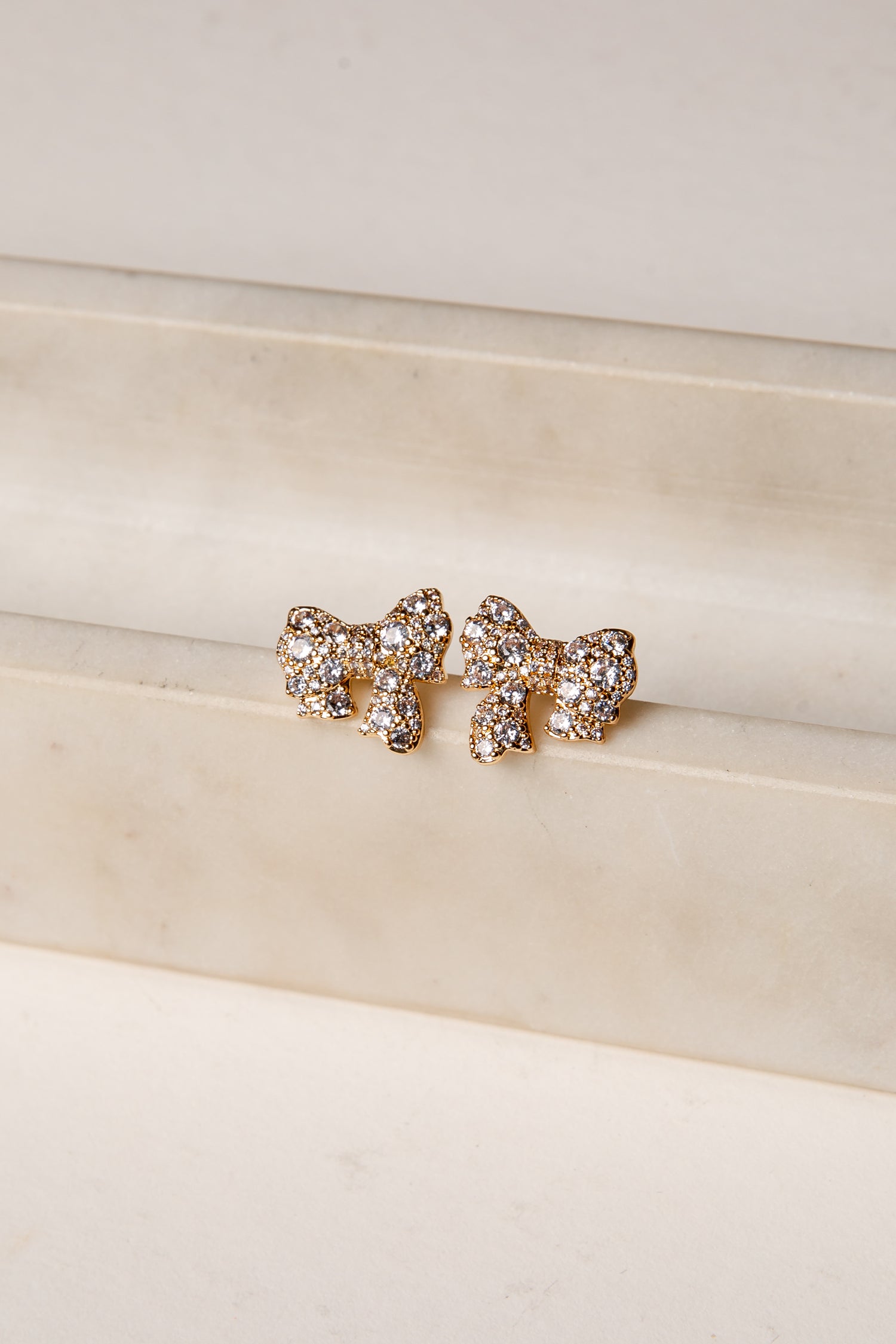 Gold bow-shaped earrings with rhinestones on a beige surface