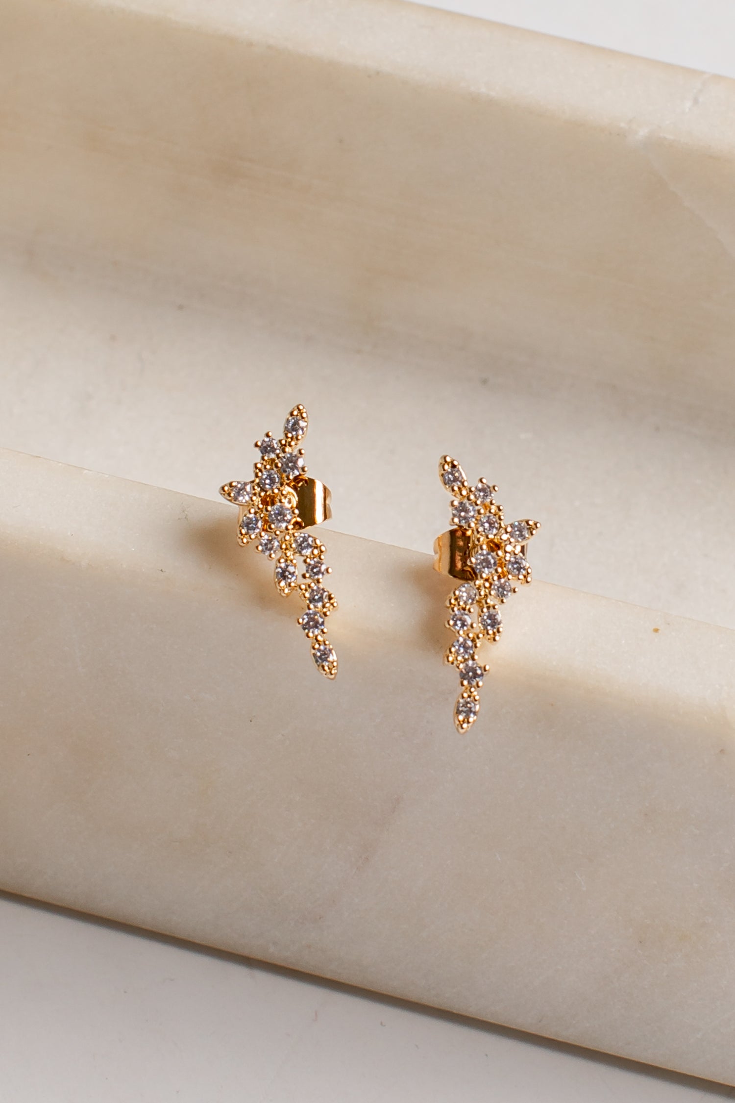 A pair of gold-toned dangle earrings with clear rhinestones, displayed on a cream-colored marble block.