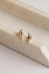 A pair of gold-toned leaf-shaped stud earrings adorned with small clear crystals, displayed on a cream-colored stone riser against a light neutral background.