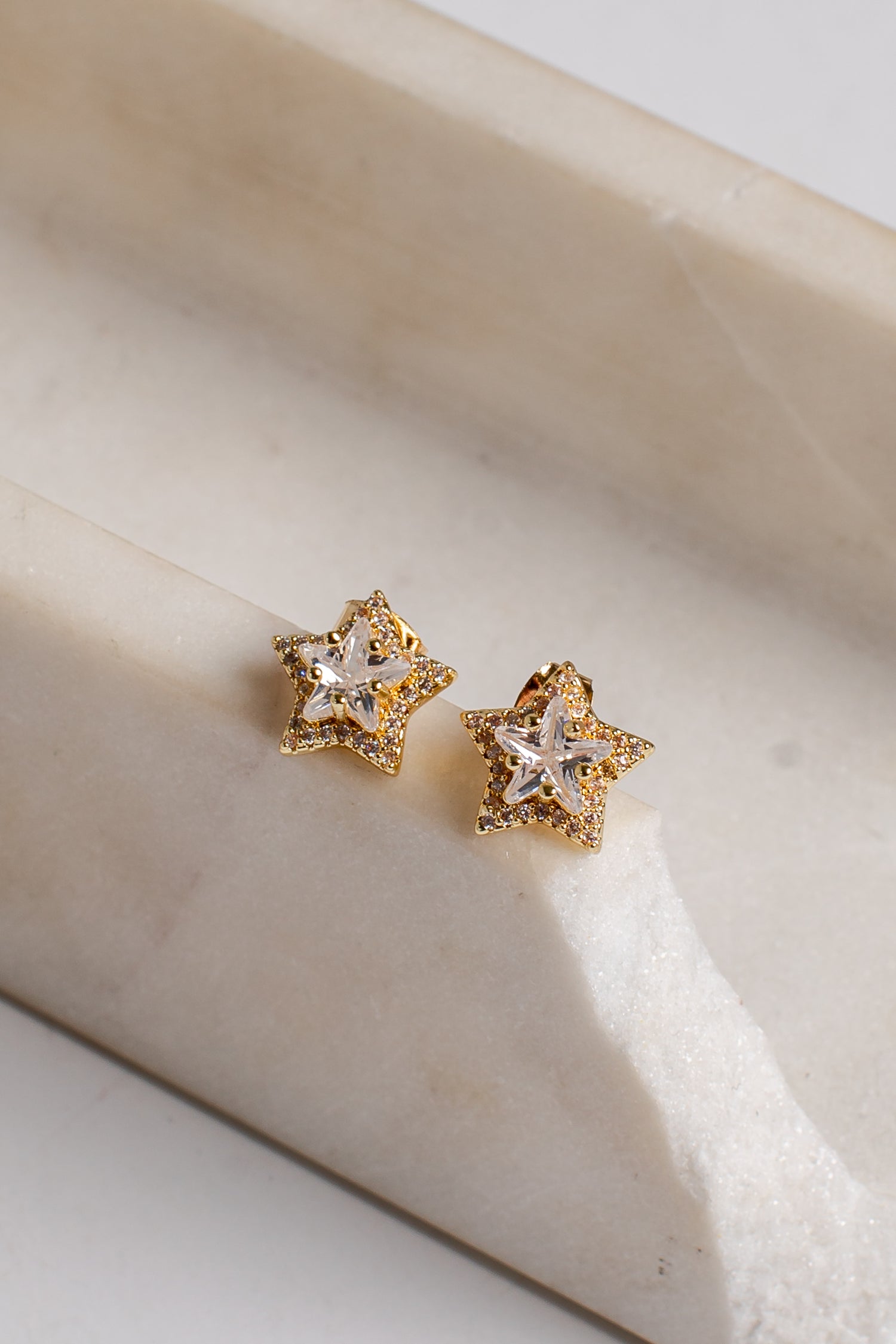A pair of gold star-shaped stud earrings with clear crystal centers, resting on a light beige marble surface.