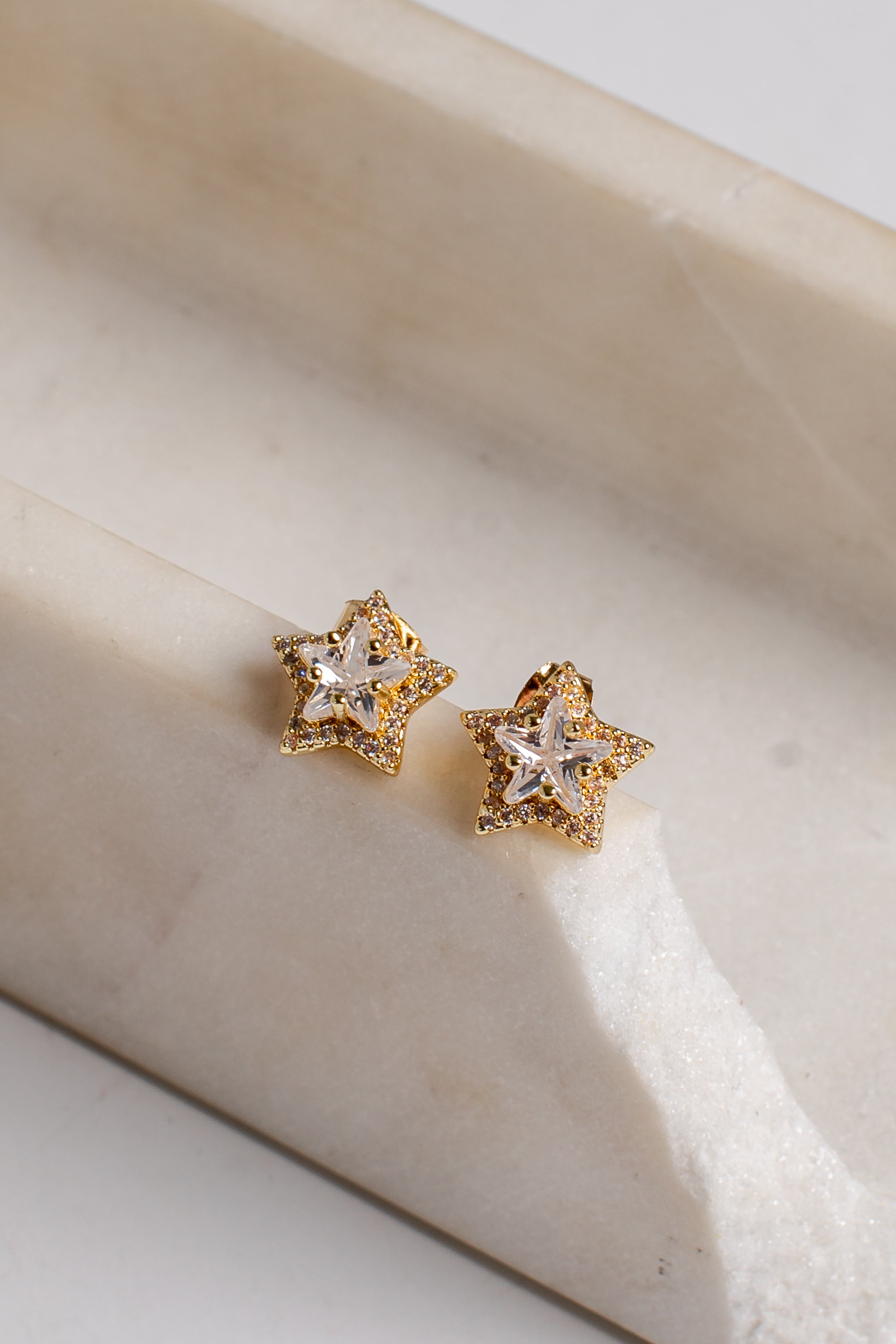 A pair of gold star-shaped stud earrings with clear crystal centers, resting on a light beige marble surface.