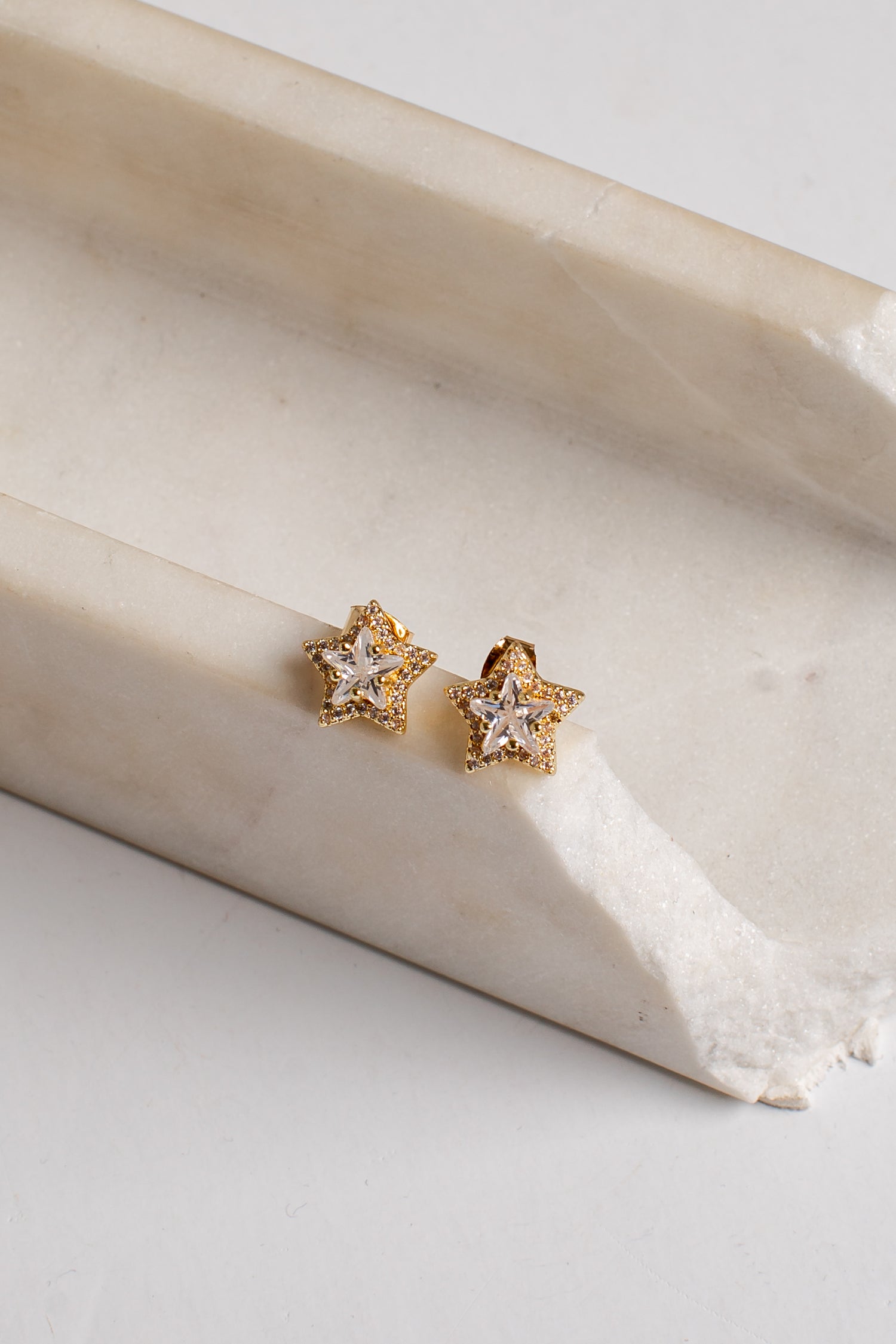 A pair of gold star-shaped stud earrings with clear gemstones, resting on a curved cream-colored stone slab against a light gray background.
