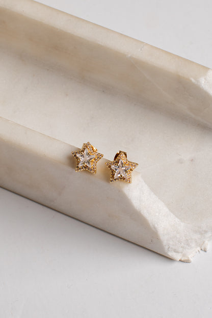 A pair of gold star-shaped stud earrings with clear gemstones, resting on a curved cream-colored stone slab against a light gray background.