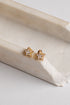 A pair of gold star-shaped stud earrings with clear gemstones, resting on a curved cream-colored stone slab against a light gray background.