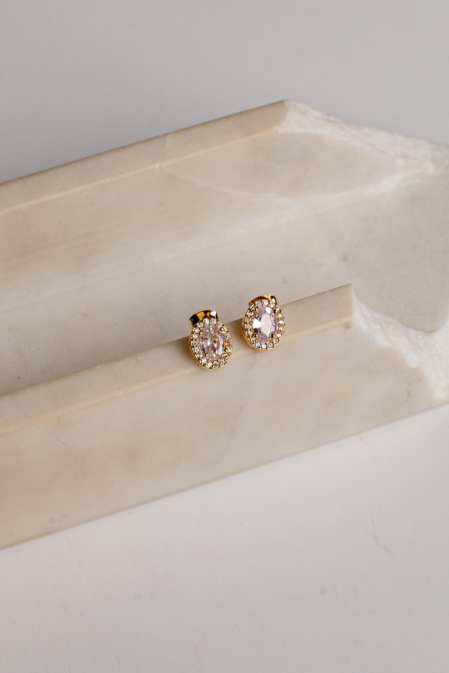 A pair of gold-toned stud earrings with oval clear gemstones and small surrounding clear stones, displayed on a cream-colored stone riser against a light neutral background.