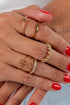 Close-up of a hand wearing multiple gold rings with red nail polish on a neutral background