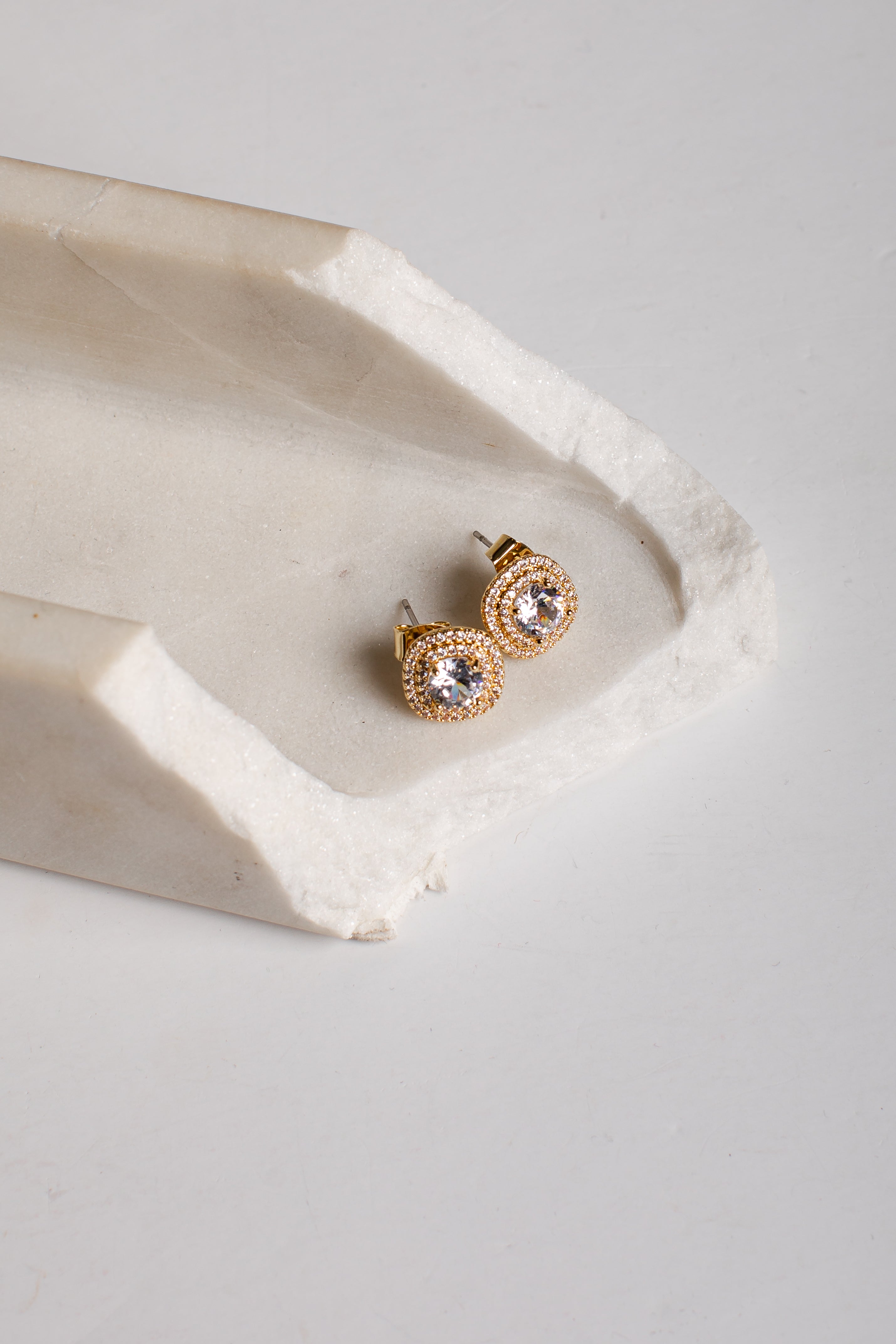 A pair of gold stud earrings with clear round gemstones, displayed on a textured white stone tray against a light gray background.