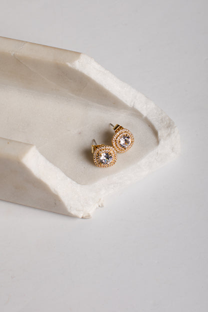 A pair of gold stud earrings with clear round gemstones, displayed on a textured white stone tray against a light gray background.