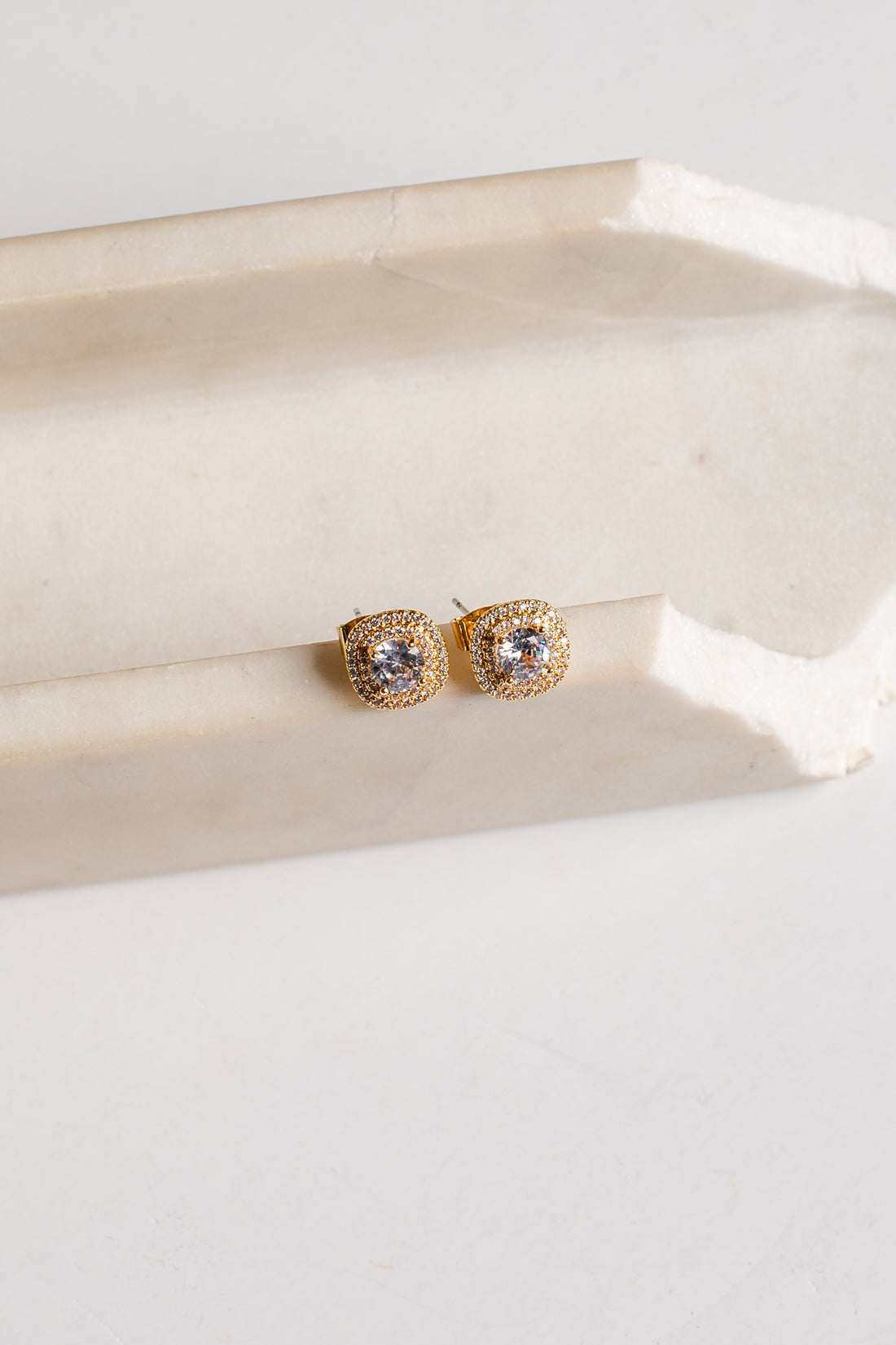 A pair of gold square stud earrings with clear stones, resting on a light beige stone block against a white background.