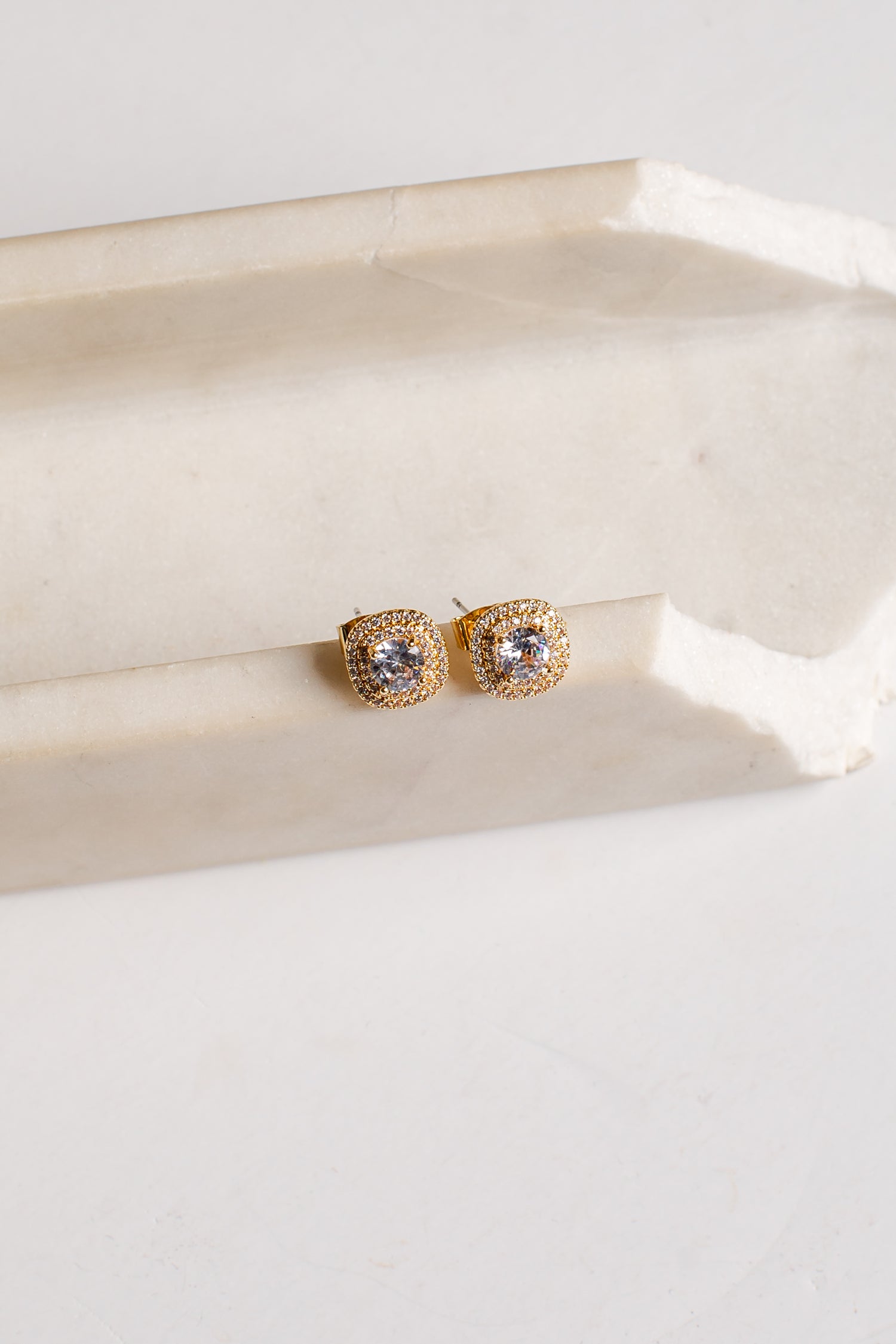 A pair of gold square stud earrings with clear stones, resting on a light beige stone block against a white background.