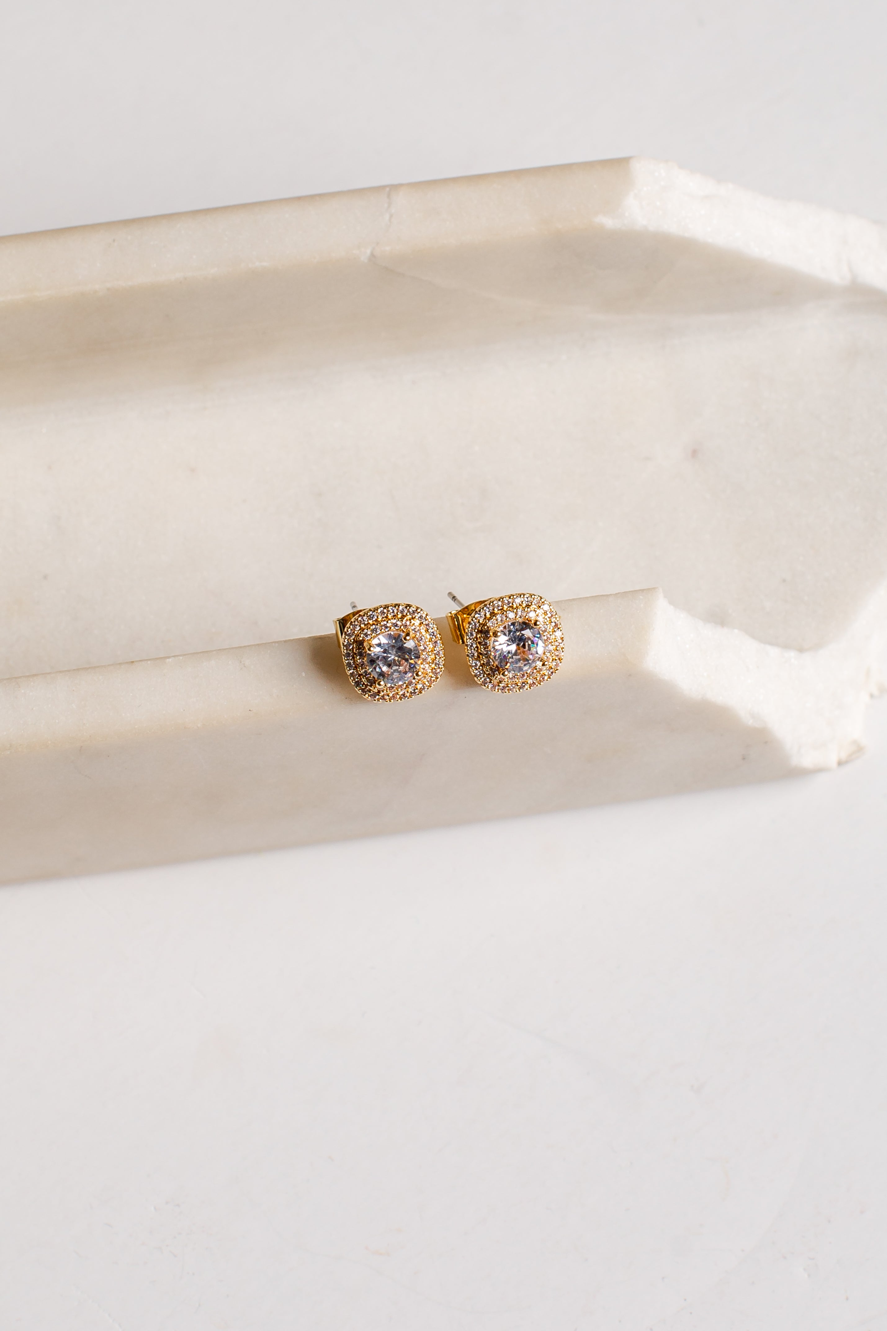 A pair of gold square stud earrings with clear stones, resting on a light beige stone block against a white background.