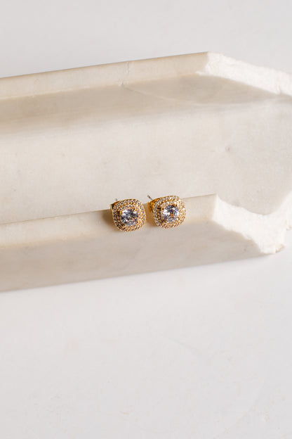 A pair of gold square stud earrings with clear stones, resting on a light beige stone block against a white background.