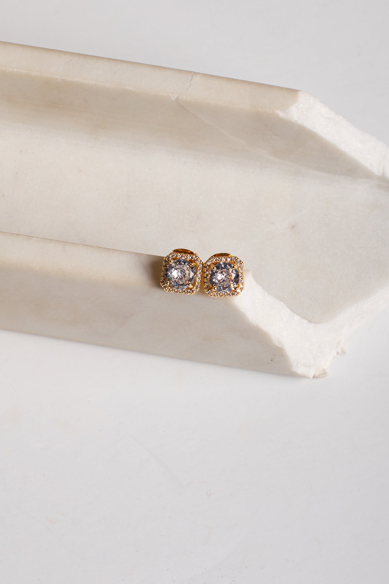 A pair of gold post earrings with clear round gemstones, resting on a light beige stone block against a white background.