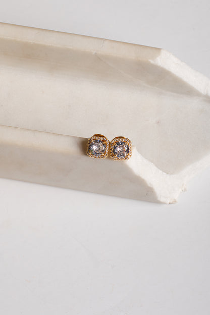 A pair of gold post earrings with clear round gemstones, resting on a light beige stone block against a white background.
