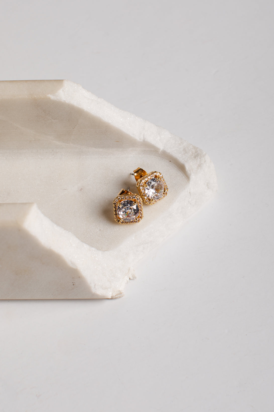 A pair of gold stud earrings with clear gemstones, resting on a textured white stone tray against a light gray background.