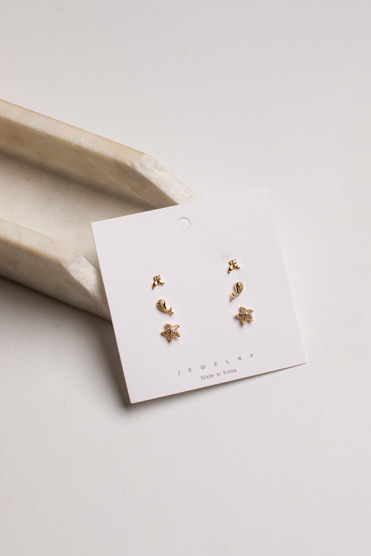A pair of small gold stud earrings displayed on a white jewelry card, photographed on a light neutral background with a beige tray prop.