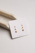 A pair of small gold stud earrings displayed on a white jewelry card, photographed on a light neutral background with a beige tray prop.