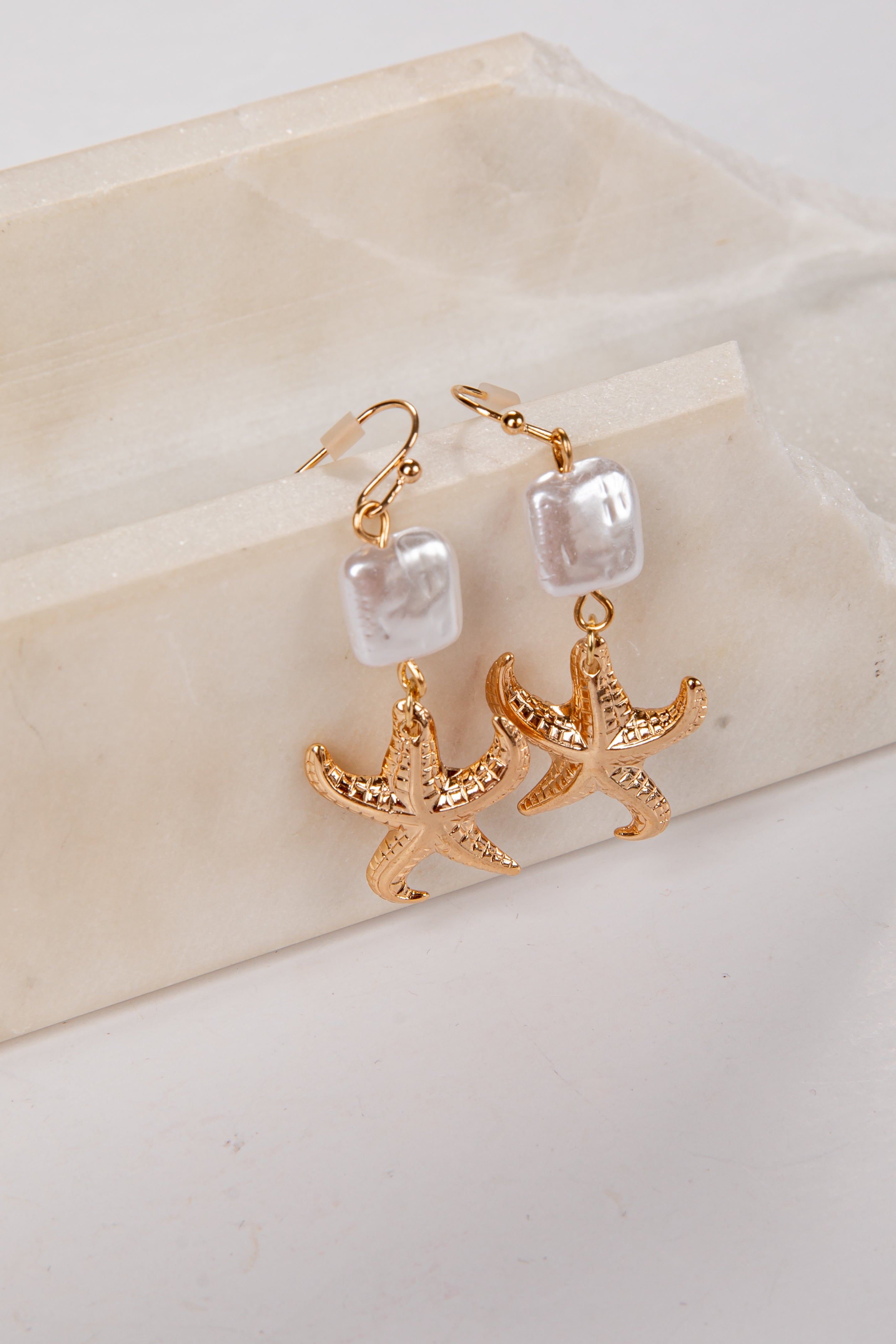 Gold starfish earrings with pearl-like beads on a beige stone surface