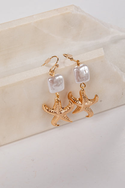 Gold starfish earrings with pearl-like beads on a beige stone surface