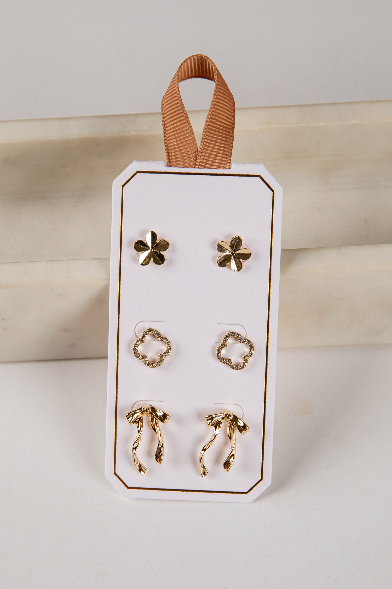 Set of gold earrings on a white card with a brown ribbon, against a neutral background.