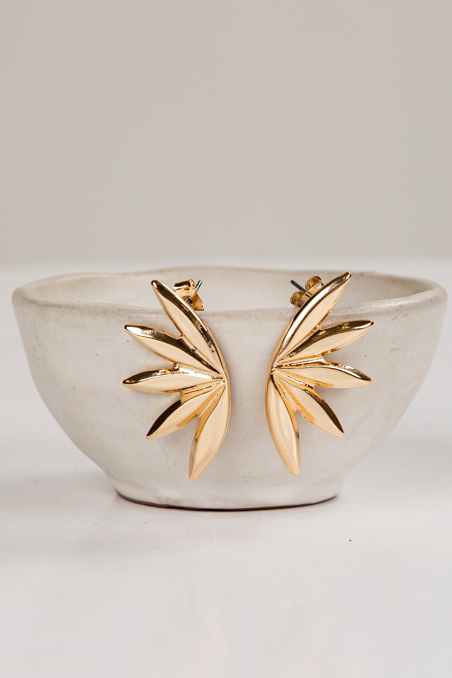 Gold leaf-shaped earrings on a white ceramic dish against a light gray background