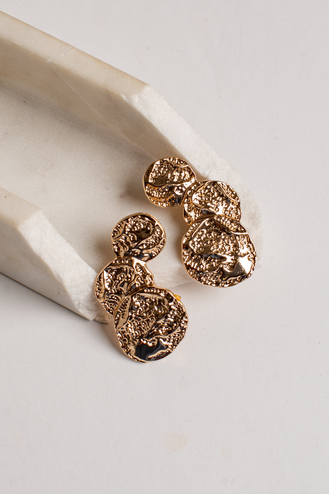 A pair of textured gold coin drop earrings resting on a white marble scoop against a light gray background.