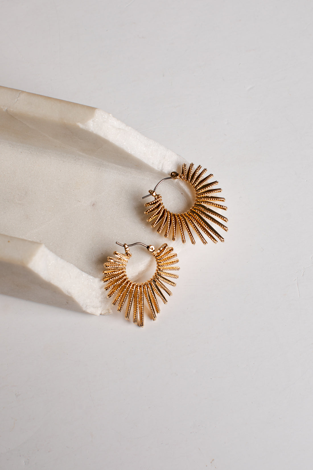 A pair of gold-toned textured hoop earrings with a sunburst or fan-like radiating design, displayed on a white marble tray against a light gray background.