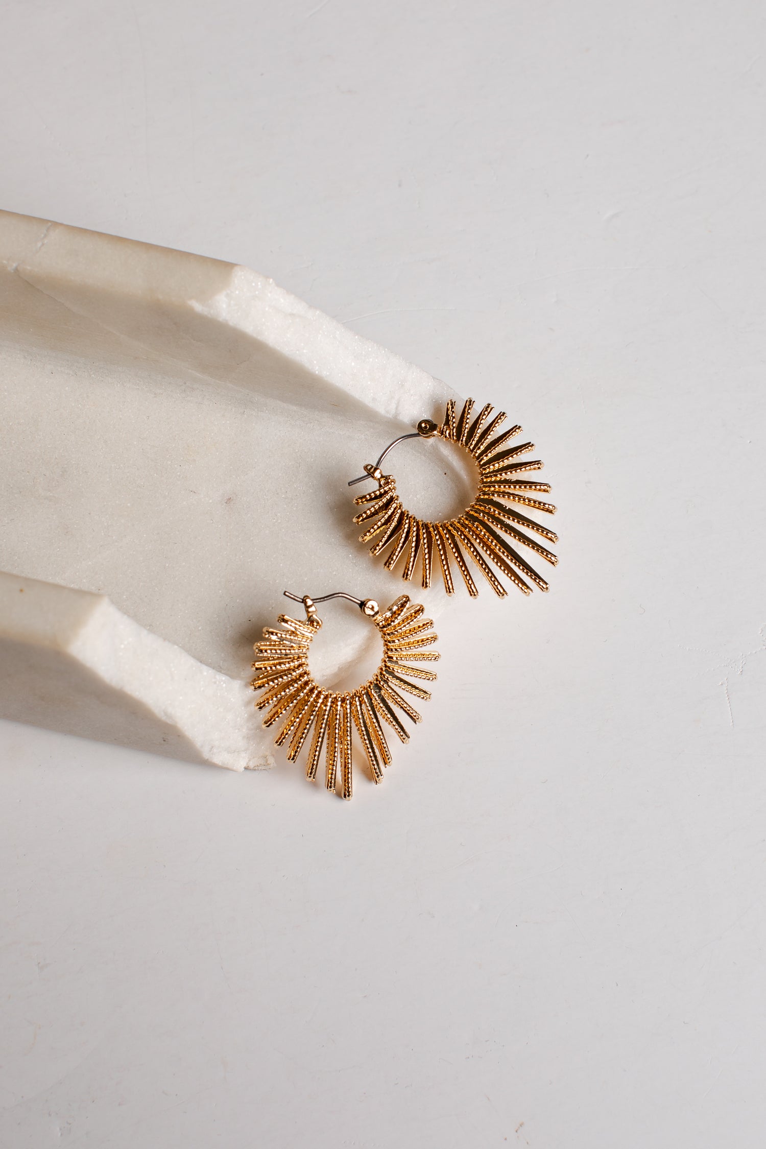 A pair of gold-toned textured hoop earrings with a sunburst or fan-like radiating design, displayed on a white marble tray against a light gray background.