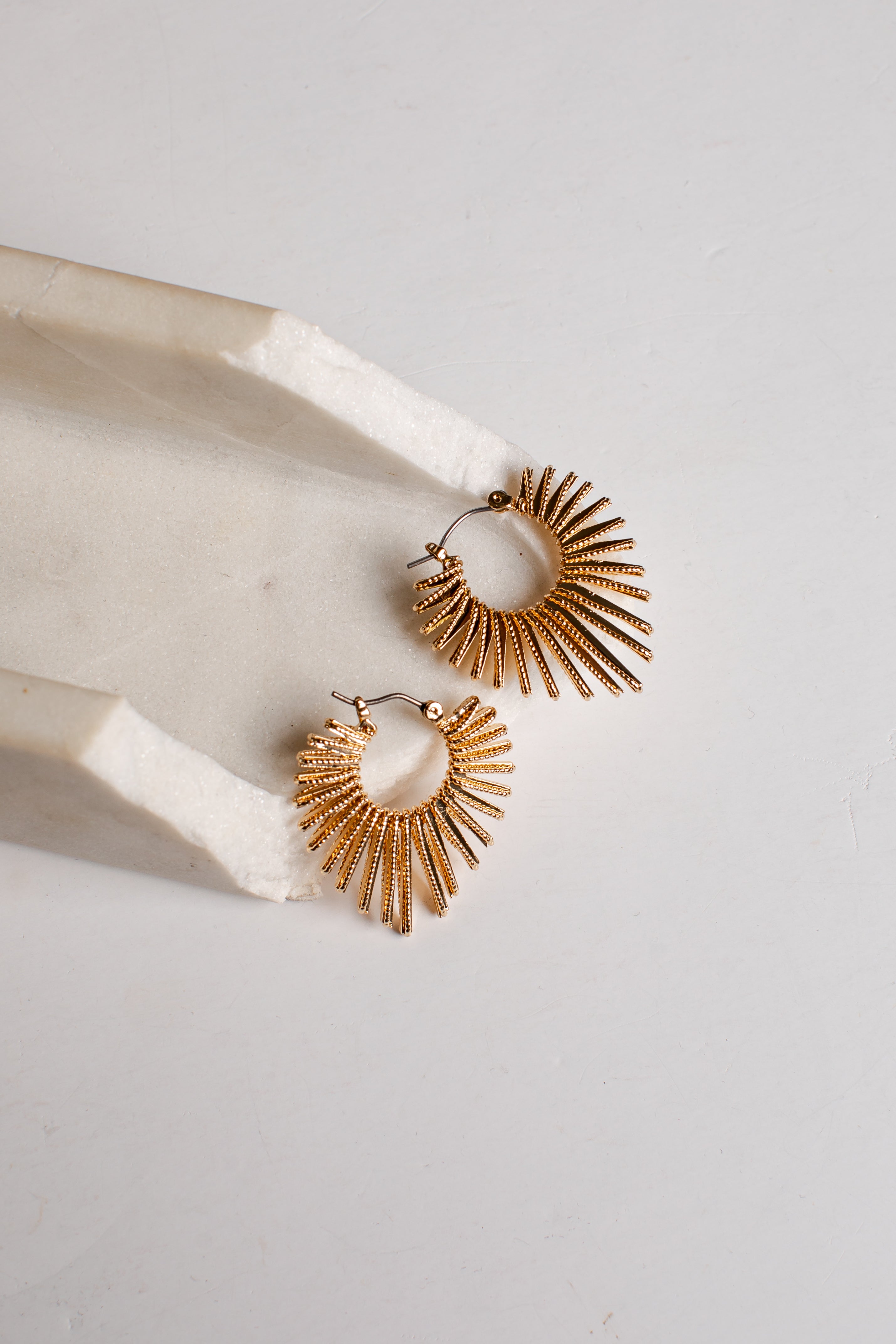 A pair of gold-toned textured hoop earrings with a sunburst or fan-like radiating design, displayed on a white marble tray against a light gray background.