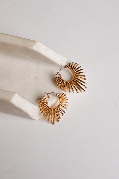 A pair of gold-toned textured hoop earrings with a sunburst or fan-like radiating design, displayed on a white marble tray against a light gray background.