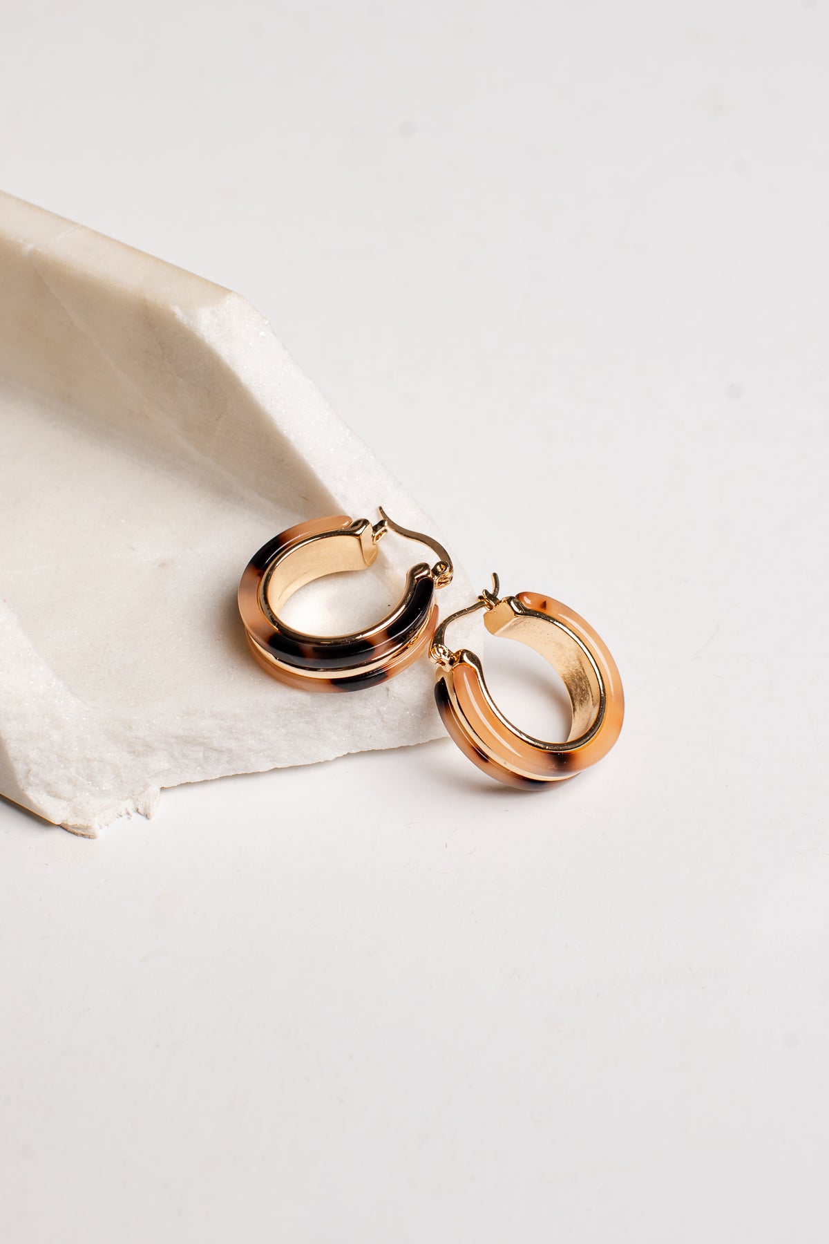 A pair of polished rose gold hoop earrings resting on a textured white stone slab against a clean white background.