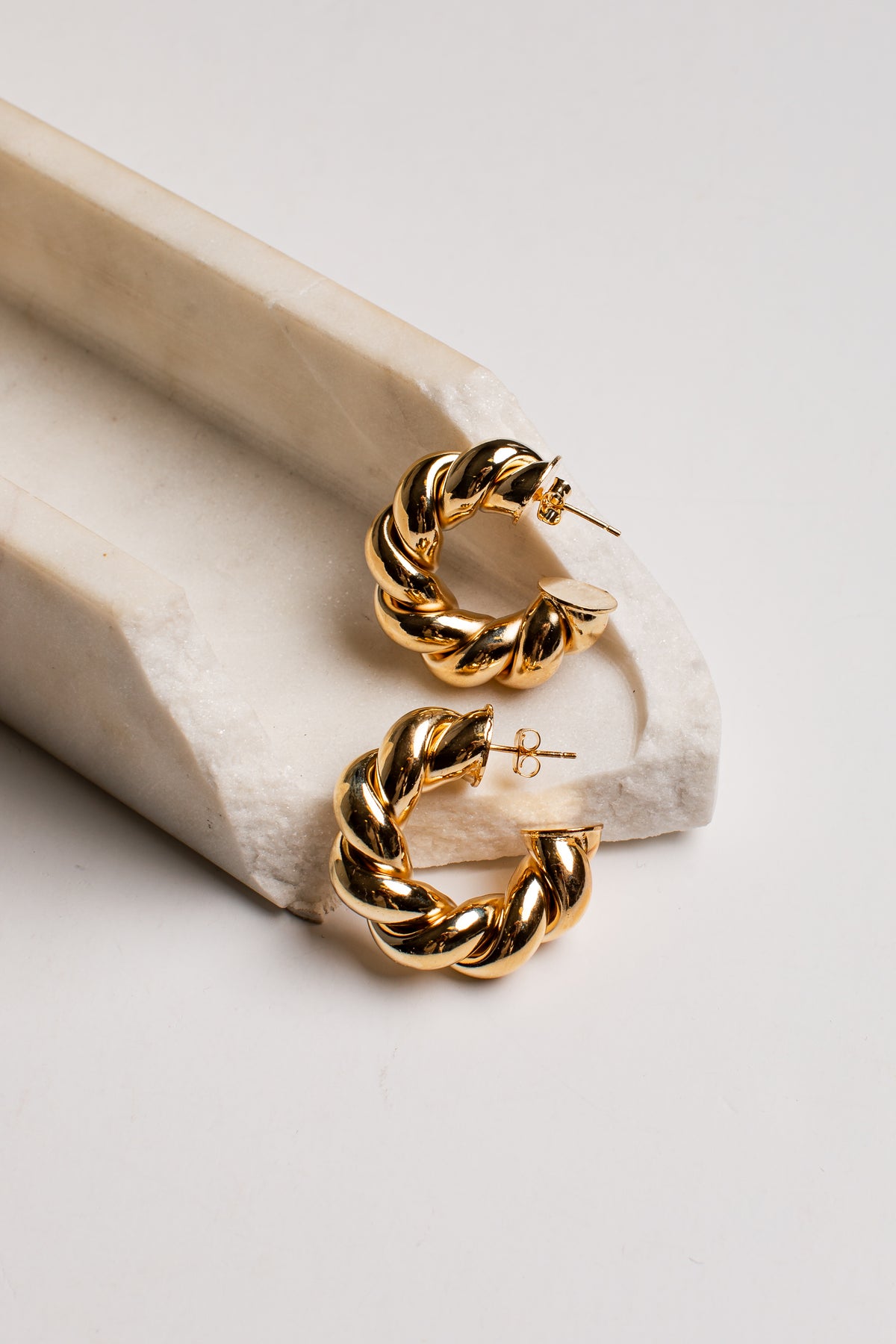 A pair of gold hoop earrings with a twisted rope texture, displayed on a light beige stone riser against a clean white background.