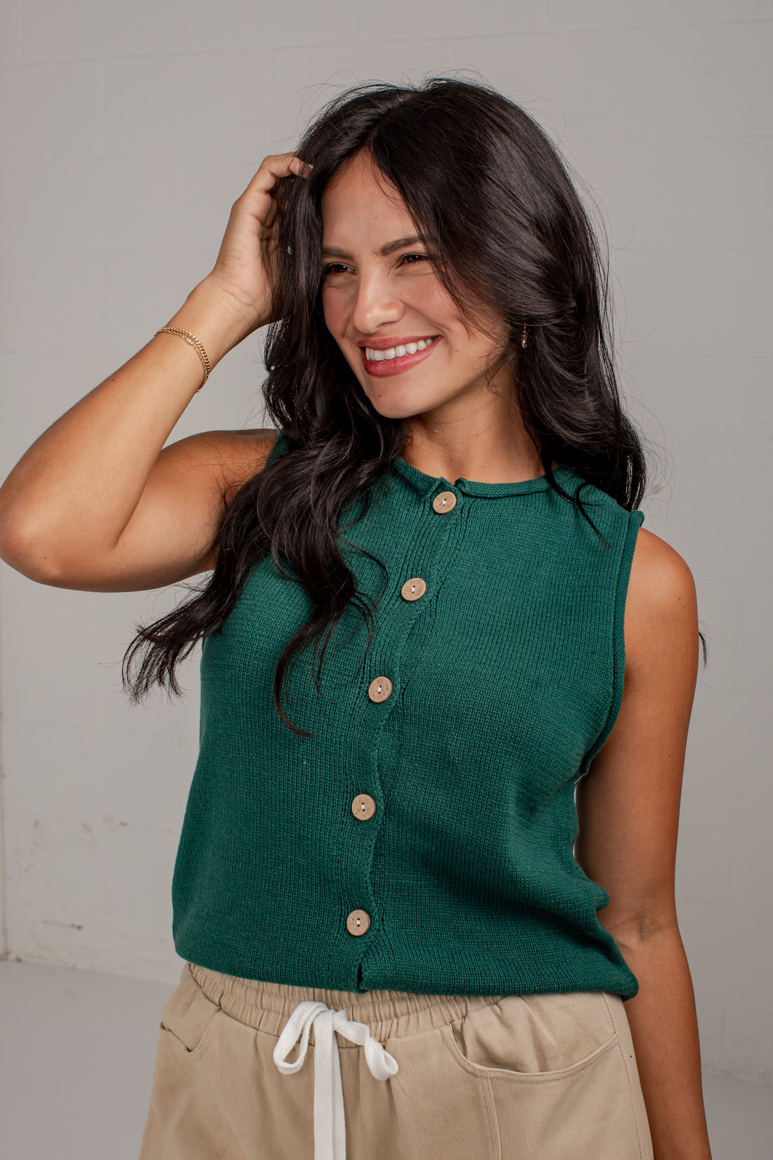A woman wearing a sleeveless dark green knit top with a button-down front and beige drawstring pants, posing against a light gray wall.