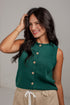 A woman wearing a sleeveless dark green knit top with a button-down front and beige drawstring pants, posing against a light gray wall.