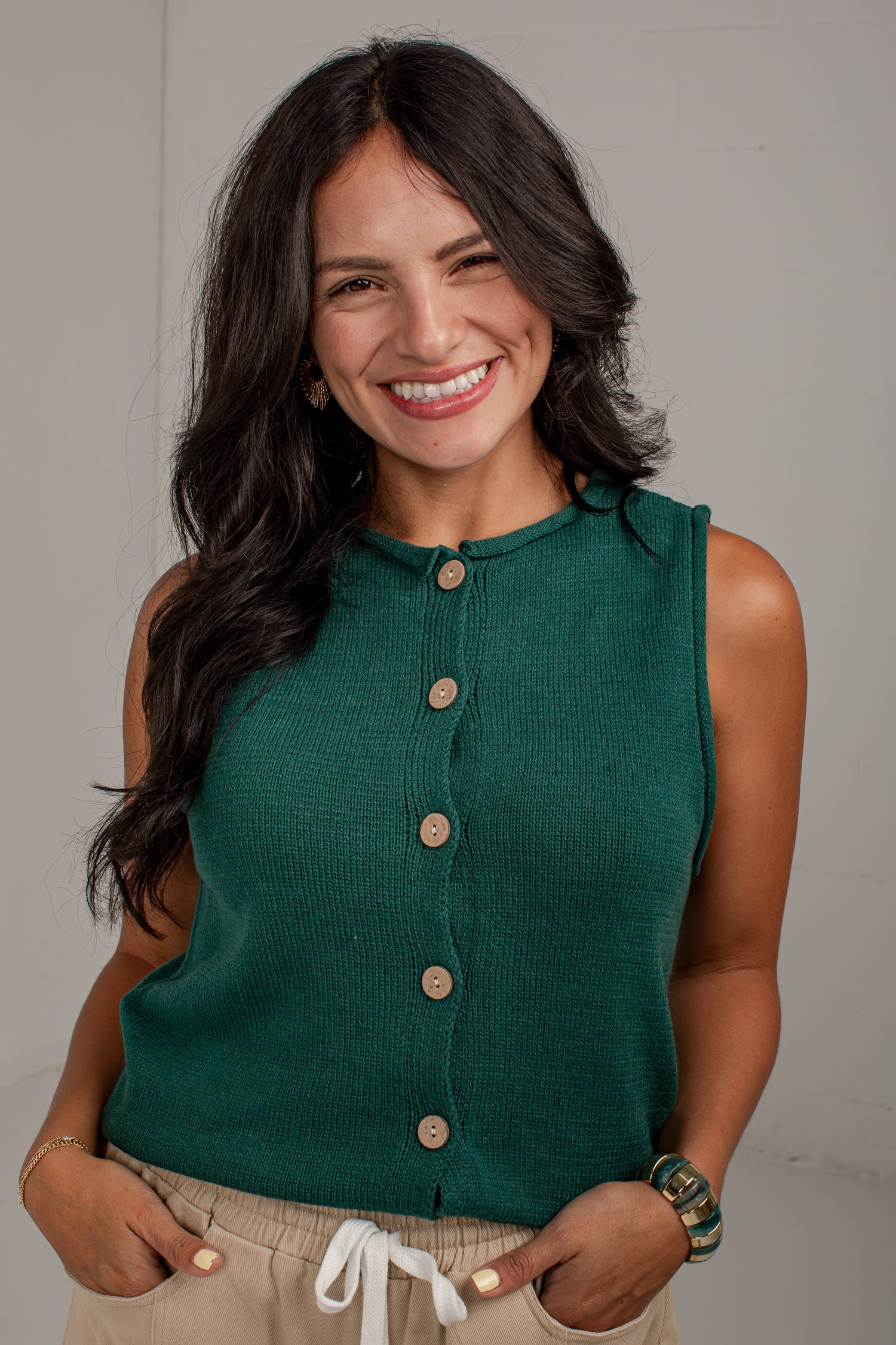 A smiling woman with long dark hair wearing a sleeveless dark green ribbed knit top with a button-down front, paired with beige drawstring pants, standing against a light gray wall.