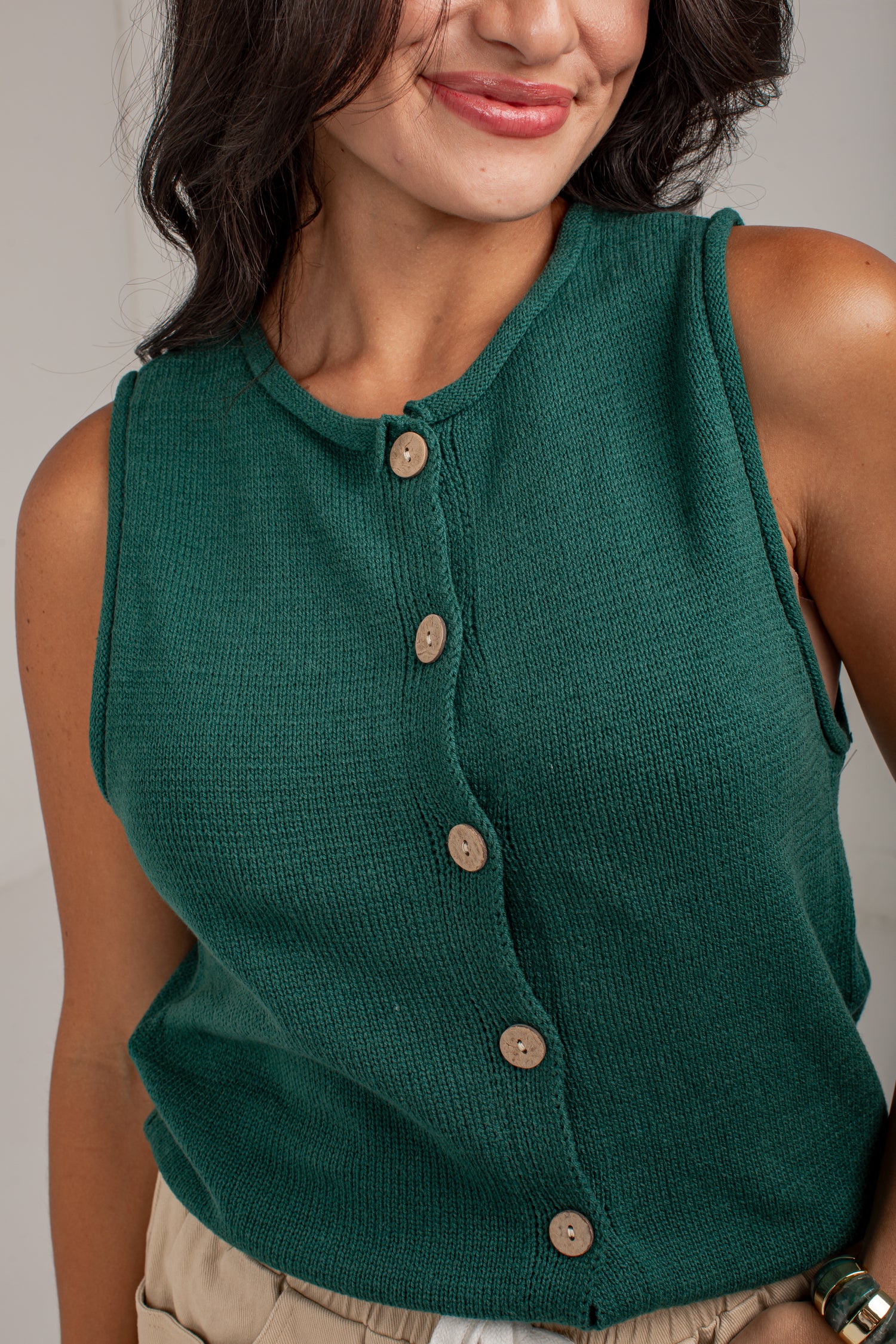 A close-up of a woman wearing a sleeveless, ribbed knit top in forest green with a button-down front. The top features light brown wooden buttons.