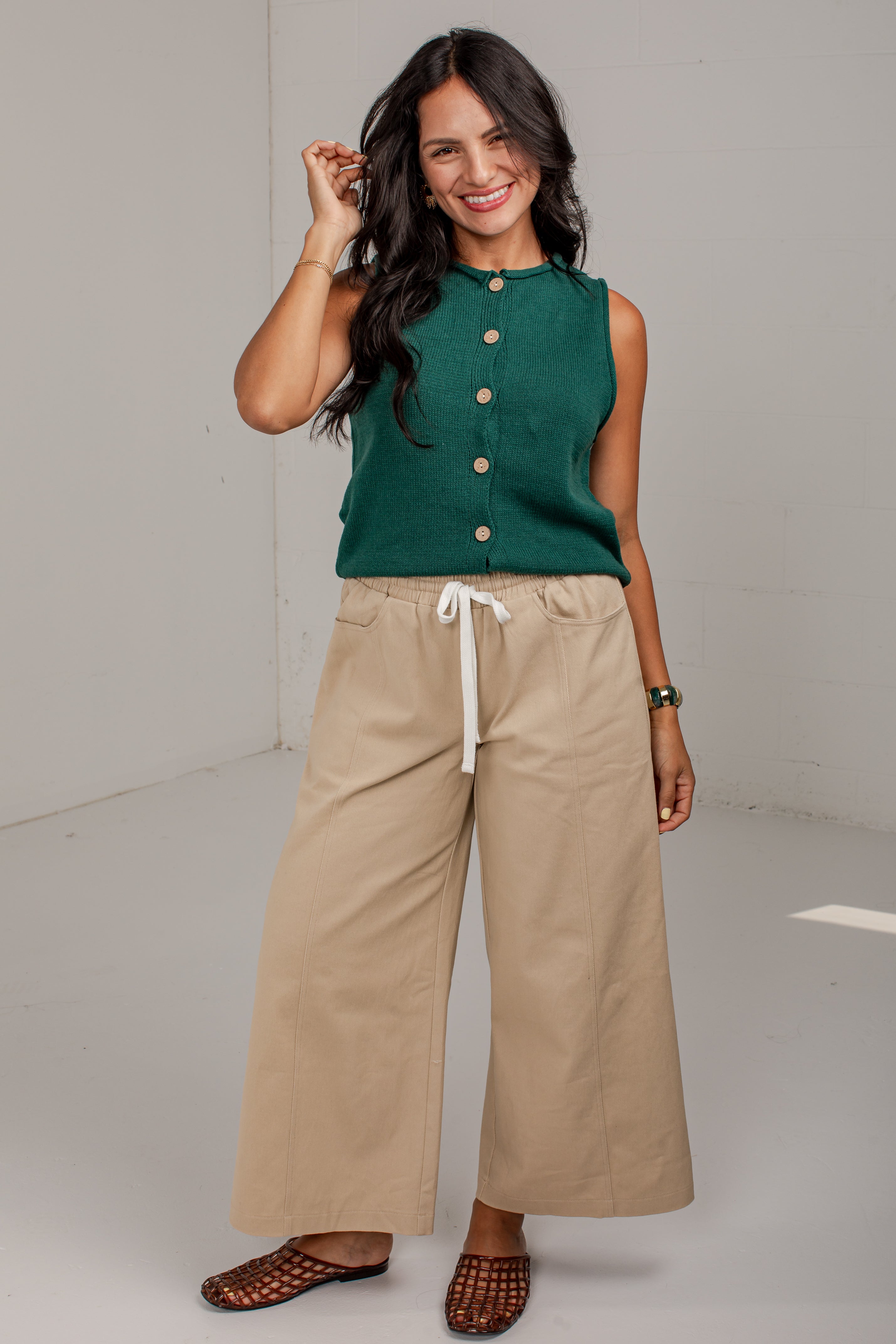 A woman modeling a sleeveless dark green knit top with gold buttons and beige wide-leg drawstring pants, paired with brown woven slide sandals, in a bright studio setting.