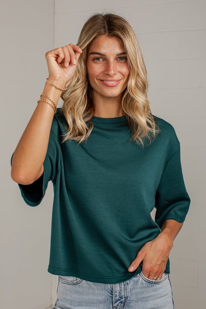 A woman with blonde hair wearing a dark green short-sleeve t-shirt and light blue jeans, standing against a light gray paneled wall.